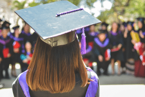  woman with her back turned wearing an academic cap while looking at her classmates who are blurry in the background