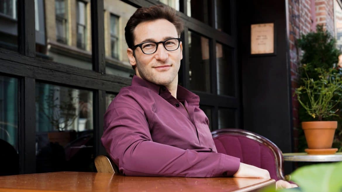 Simon Sinek in a maroon polo shirt with one hand on a table, smiling