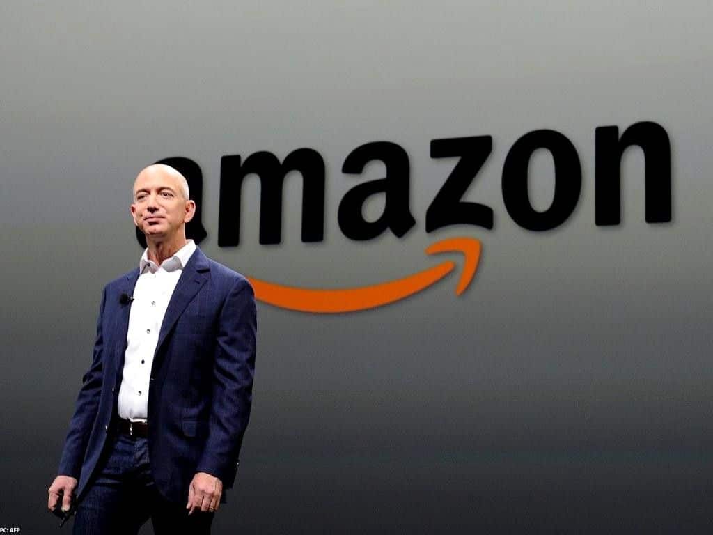 Standing Jeff Bezos on a blue suit with Amazon’s logo behind him