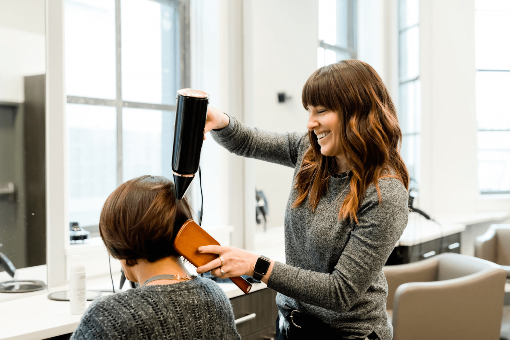 smiling woman holding a hairdryer over another woman’s hair, with her other hand holding a brush