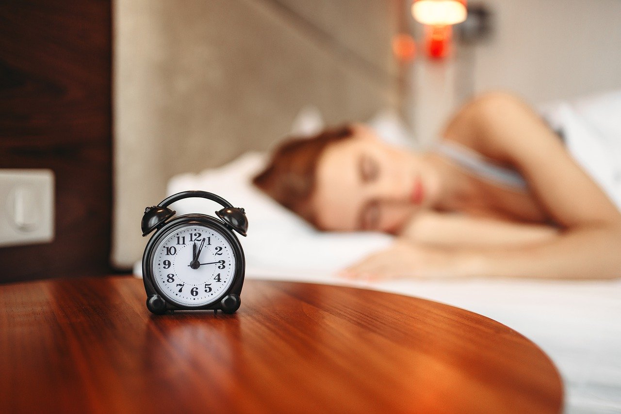 A black alarm clock on a wooden table beside a sleeping woman