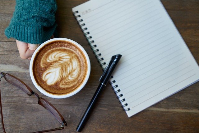 A notebook, pen, and glasses near a coffee cup held by a person