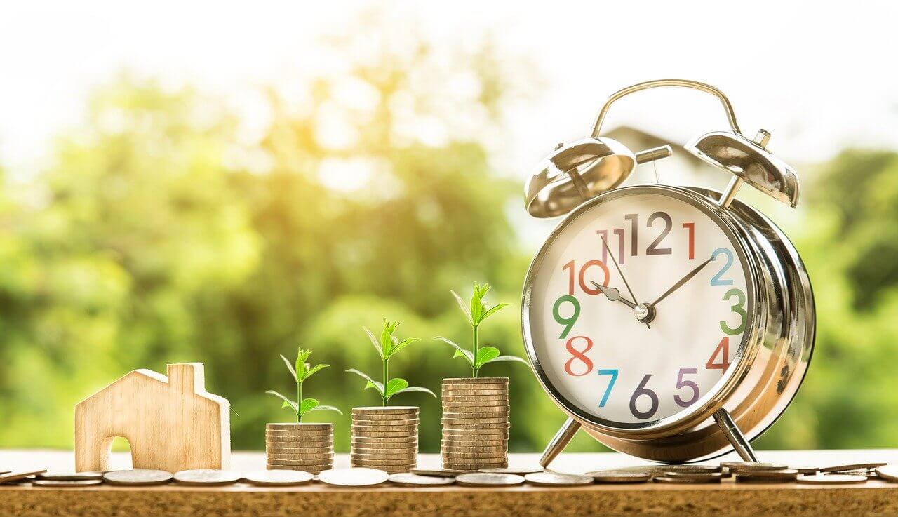 A wooden house outline and a silver alarm clock beside 3 stacks of coins with sprouts on top of them