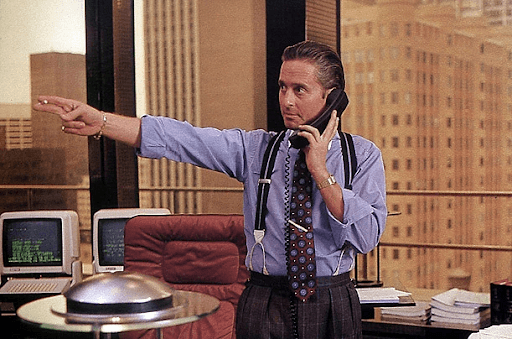  Gordon Gekko on a blue long-sleeve polo talking to someone on the phone while pointing to his left side