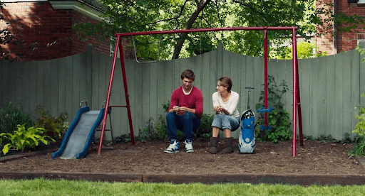 Gus and Hazel talking while sitting on a kid’s swing