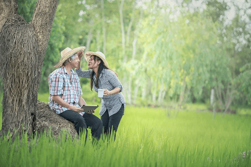  A woman holding a cup and an old man seated near a tree trunk on a grassy countryside