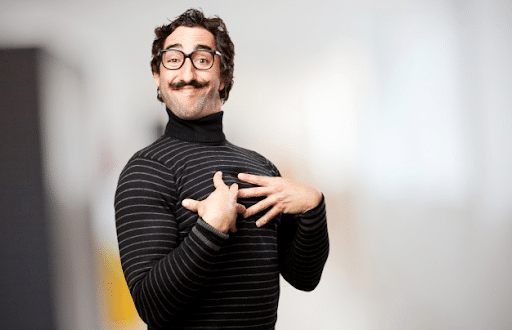 A bearded man with glasses wearing a black turtleneck long sleeve blouse posing his hands on his chest