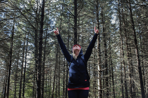 A woman in a forest wearing a dark blue hoodie raising her hands up