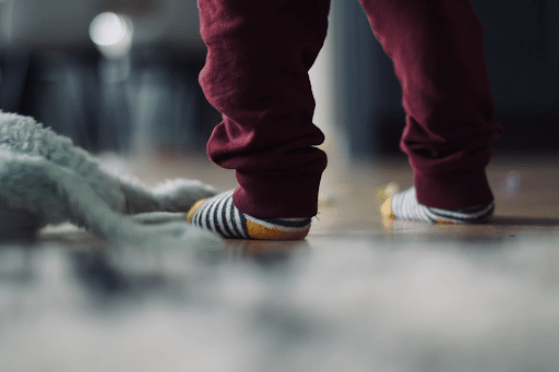 Legs of a toddler wearing red pants and socks standing next to a doll on the floor