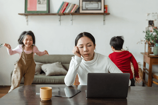  kids running in the background while mom is tired and facing her laptop