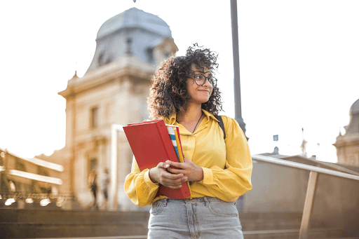 A curly-haired female student in a yellow long-sleeve polo shirt holding books