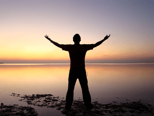 Silhouette of a man extending his arms up while standing beside an ocean during sunset