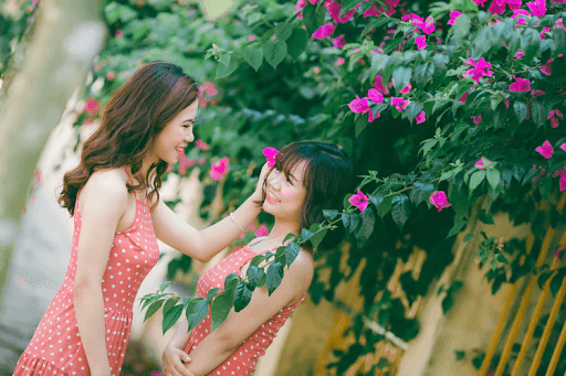 Two young women in dotted, sleeveless shirts beside flowers and smiling at each other