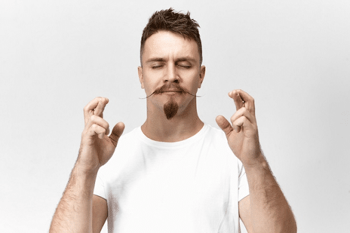 Young bearded hipster guy with closed eyes and crossed fingers while wishing for good luck