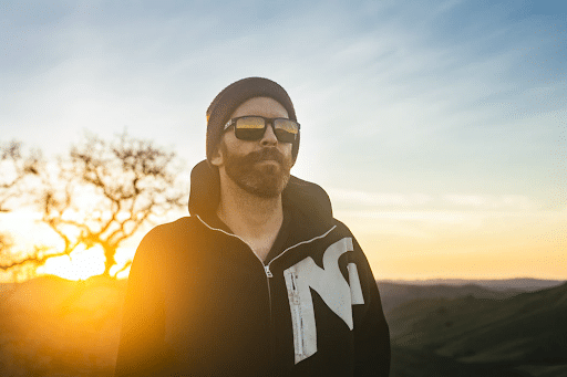 cool man wearing sunglasses and a hoodie looking off into the distance on a mountain overlooking a hill landscape at sunset