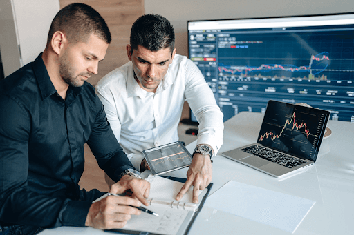 Two men in white and black long-sleeve polos in front of a laptop with a graph displayed